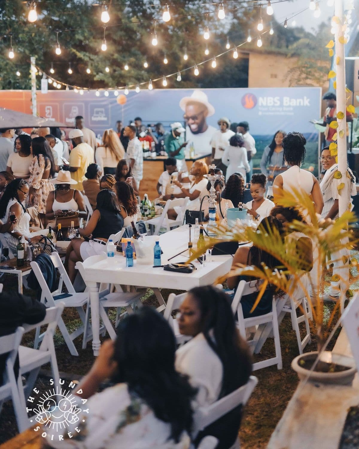 Soirée d'entreprise en extérieur — tables blanches, guirlandes, groupe réuni à la tombée de la nuit.
